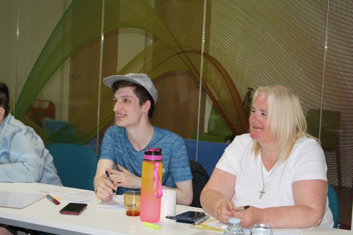 Young man with baseball cap and lady with blonde hair sit together in classroom looking at tutor